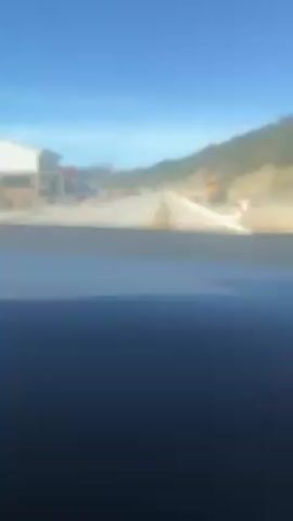 The video shows a car driving on a paved road on a sunny day. There is construction on the side of the road, with a yellow bulldozer visible. A black Volkswagen Tiguan SUV passes in the opposite direction. Buildings are on the left side of the road, and mountains are visible in the distance. The car's dashboard is visible in the foreground. Music plays in the background.
