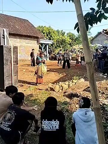 The video shows a group of people gathered in what appears to be a village or rural area. A person dressed in traditional attire stands near a large speaker, with other people in similar clothing walking around in the background. Some onlookers are seated in the foreground. The setting includes brick buildings, trees, and a dirt ground with rocks. The sky is clear and blue, indicating it's a sunny day.