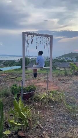 A person sits on a swing facing away from the camera, looking out at a landscape. The swing set has decorative elements hanging from it. The scene is outdoors with vegetation in the foreground and a town or village in the background. The sky is cloudy with a hint of sunset colors. Further in the distance, other people are seated at tables, also looking out at the view.