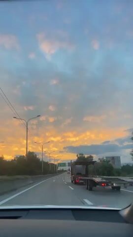 The video shows a car's point-of-view driving on a highway at sunset. The sky is filled with dramatic clouds in shades of orange, pink, and blue, indicating either sunrise or sunset. The setting appears to be a rural or semi-rural area, with trees lining the highway and occasional buildings visible in the distance.

The highway has multiple lanes, and traffic is moderate, with a few cars and trucks. A car carrier truck is prominently visible in the right lane, moving in the same direction as the camera. Overhead streetlights are spaced along the road.

There are several road signs visible. One green sign indicates directions to "E60," "BATUMI," "TBILISI," and "SUKHUMI" with arrows pointing forward and to the right. Another blue sign indicates directions to "KASANI," "AKHALGORI," and has a sign for a service area with a car and a bed icon. The text on the signs is in both Georgian and English.

The overall mood is serene and contemplative, enhanced by the beautiful sunset and the smooth movement of the car. The time of day is clearly sunset, with the warm hues of the sky casting a glow over the scene. The location is identified as Mukhrani, Georgia, through the road signs. There are no people directly visible outside of the vehicles, but the presence of vehicles suggests ongoing travel.