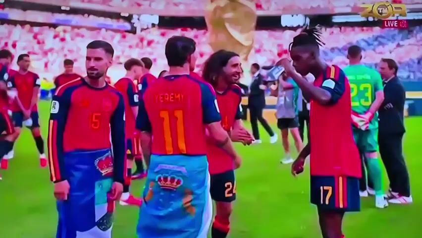 The video shows a group of male soccer players in red jerseys on a green field. In the foreground, a player with the number 17 on his jersey is drinking from a bottle. To his left, a player with the number 11 on his jersey, identified as JEREMY, is talking to him and gesturing with his hands. Behind them, other players in red jerseys are visible, some with numbers on their backs, including 15 and 25. Some players are holding flags with emblems. In the background, a large gold trophy is visible, and a crowd of spectators can be seen in the stands. The scene appears to be a post-game celebration or gathering. The broadcast information in the upper right corner indicates "ZOR LIVE".