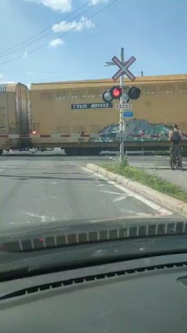 A yellow freight train is passing through a railroad crossing. A person is on a bicycle to the right of the crossing. The crossing gate is down, indicated by the red lights flashing. The train cars are labeled TTUX and CN. The weather appears to be sunny with some clouds. The camera is inside a vehicle, looking out at the scene.