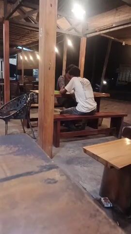 The scene captures a nocturnal view of what appears to be an open-air or semi-covered cafe or restaurant in Geulumpangmeujim-Jim, Indonesia. Artificial lighting, primarily from bare bulbs hanging from the ceiling structure, illuminates the space.

The establishment features a rustic aesthetic, characterized by sturdy wooden pillars, exposed wooden beams forming the roof, and a concrete floor. Numerous wooden tables, some paired with long wooden benches, are arranged throughout the area. Several black metal chairs with intricate patterns are also visible, one prominently placed in the mid-ground.

In the mid-ground, two men are seated at one of the wooden tables with benches. One man, wearing a white t-shirt with a design on the back, has his back to the camera. The other man, dressed in a darker long-sleeved shirt, faces him. They appear to be engaged in a casual interaction or conversation, as the man in the white shirt briefly stands up and then reseats himself.

In the immediate foreground, a wooden tabletop, slightly out of focus, occupies the bottom left of the frame. To the right, another smaller wooden table holds a white rectangular object, possibly a cigarette pack or matchbox. The background is dark, suggesting the area is outdoors, with faint lights visible in the distance. A clear numerical label "3" is affixed to one of the prominent wooden pillars, possibly indicating a section or table number. The overall atmosphere appears relaxed and casual.