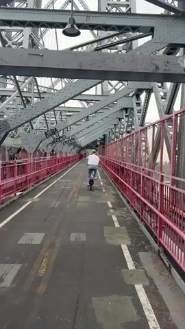 This media file captures a POV perspective of a person riding a bicycle across a bridge in New York City. The bridge is characterized by its prominent gray metal truss structure and a distinctive bright pink railing that lines the pedestrian and bike path. The path itself is paved and shows signs of wear, with yellow and white lane markers and some graffiti.

In the foreground, a person on a bicycle is visible, riding away from the camera. They are wearing a white t-shirt and shorts, and the bicycle appears to be a small, possibly electric, model. Further ahead on the bridge, another individual on a bicycle can be seen.

The scene is set on a bridge, likely a pedestrian and cyclist walkway, offering views of a body of water and the cityscape in the distance. The weather appears to be overcast, with a grayish sky, suggesting it might be morning or late afternoon. The time of day is not explicitly stated but the lighting indicates it's not the middle of the night.

Notable details include the intricate metalwork of the bridge's superstructure, the vibrant pink railings, and the presence of graffiti on the bridge's surface and some of the metal components. The background shows a hazy skyline with buildings and some trees lining the waterfront on the opposite shore. The overall mood is one of urban exploration and enjoying a bike ride over a significant piece of infrastructure.

Visible text includes graffiti on the bridge structure, appearing as various tags and markings, such as "KUDA" and "BOOM". There is also a small white sign on the pink railing that is too blurry to read.