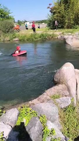 In this video, a person wearing a red helmet and a dark-colored wetsuit is riding a red raft or board down a concrete spillway into a river. The spillway creates large waves as the person navigates the turbulent water. The river is surrounded by green vegetation, trees, and concrete barriers. There are other people visible in the background near the starting point of the spillway. The sky is clear and blue.