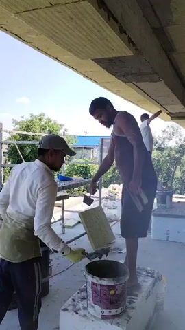 Two men are working on a construction site under a concrete overhang. The man on the left, wearing a white shirt, dark pants, and a green apron, is holding a trowel and a bucket of what appears to be mortar. He is wearing gloves and a baseball cap. The man on the right, wearing a tank top and dark shorts, is standing on a white block, also holding a trowel and applying material to the underside of the overhang. His back is mostly to the camera. Further in the background, another man in a white shirt can be seen working on the overhang.

The scene appears to be outdoors during the day, with bright sunlight casting shadows and highlighting the work. Green trees and a glimpse of a blue roof are visible behind the construction area, suggesting a rural or suburban setting. The overall impression is one of manual labor and construction. There is no discernible text visible in the frame.