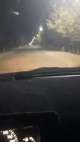 The video shows a view from the interior of a car at night. The dashboard is visible in the foreground, with a speedometer showing a speed between 140 and 160, likely kilometers per hour given the context. The road ahead is illuminated by streetlights and the car's headlights, creating a hazy, orange glow. Trees line both sides of the road, their leaves appearing somewhat blurred due to the movement of the car. In the distance, streetlights create a path of light leading into the darkness. The overall impression is of driving at night on a tree-lined road.