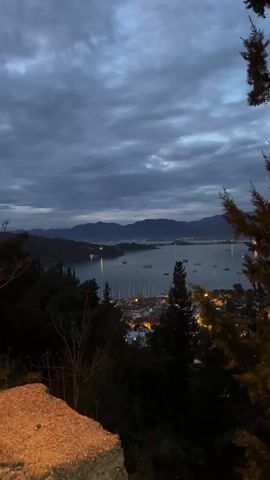 The video shows a landscape view of a bay or harbor at dusk or dawn. The sky is covered with dark, cloudy formations, giving the impression of an overcast day. In the distance, there are dark hills or mountains. The water is calm, reflecting the muted light from the sky and some scattered lights on the far shore.  

In the middle ground, a town or village is visible, with buildings and streetlights illuminating the scene. A marina with sailboats is also visible.  

In the foreground, there is greenery including pine trees, partially obscuring the view, with a stone wall along the bottom left.