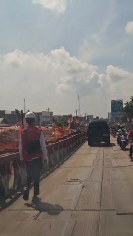 A short video clip shows a person in a white hard hat and a red vest walking across a bridge. The bridge appears to be under construction, with orange safety netting along the sides. There are Indonesian flags lining the bridge. Several motorcycles and a black van are also on the bridge, with traffic moving in the same direction as the person walking. The sky is cloudy with patches of blue.