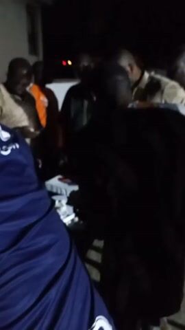 A group of about five men are gathered outside a building at night. The men are of African descent. One man is wearing a blue jacket with white writing on the back. Several of the other men are wearing sweaters or jackets. The scene is dark and the video is somewhat blurry.