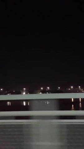 The video shows a nighttime view from a moving vehicle. The foreground consists of a white barrier, likely a guardrail, and a dark, textured surface below it. In the middle ground, a body of water reflects distant lights. The background is a dark expanse of sky above a dimly lit shoreline with scattered lights, suggesting a town or city. The overall impression is one of passing through a dark landscape illuminated by artificial lights.