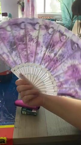 A hand holding a purple and white floral folding fan moves from right to left, obscuring a table laden with drinks and other items. The fan is partially open, with the white ribs clearly visible. In the background, a window is visible with a patterned curtain, and shelves with various objects. The lighting appears to be natural, coming from the window.