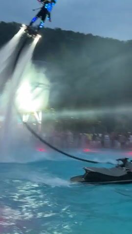 A person is jet-skiing while a person on a jetpack flies in the air, spraying water from their jetpack and propelling themselves upwards. A crowd of people is watching from the side of a pool or body of water. There are trees and a hill in the background. There are also lights around the pool. The sky is dark and cloudy, suggesting it is either dawn or dusk.