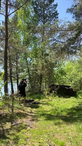 A man walks slowly towards the camera in a sun-drenched forested area. He is wearing a dark hooded sweatshirt, dark shorts, and light-colored shoes, with sunglasses obscuring his eyes. Tall pine trees and other deciduous trees surround him, their leaves vibrant green under a clear blue sky. To the left, a body of water, likely a lake or river, is visible through the trees. On the right, a large dark rock or tree stump sits on the grassy ground. The ground is a mix of dirt paths and green grass, with shadows from the trees dappling the scene. The overall impression is a peaceful outdoor setting in Sliznevo, Russia.