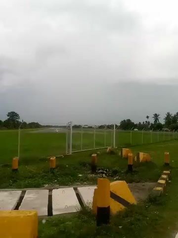 The video shows a view from a moving vehicle on a paved road. A grassy field bordered by a chain-link fence is on the left side of the road. Beyond the fence, there is an open area, possibly an airfield, with some structures in the distance. Palm trees and other vegetation line the right side of the road, with some buildings visible amongst them. Several cars are driving on the road ahead of the camera. The sky is overcast with gray clouds. The overall impression is a somewhat overcast day in a rural or semi-rural area.