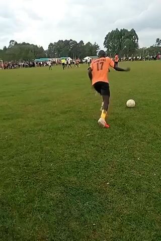 The video shows a soccer game in progress. A man wearing a black and orange jersey with the number 17 is facing away from the camera, in the foreground, on a grassy field. In the background, other players can be seen running around the field during the game. A crowd of people is standing on the sidelines. The sky is overcast with trees lining the edge of the field.