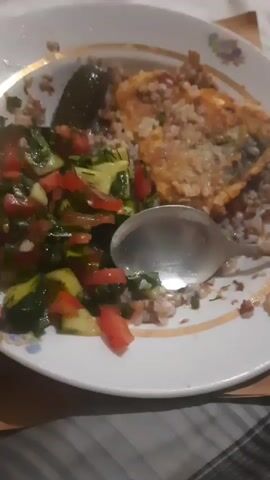A plate of food sits on a table. The plate contains a portion of buckwheat, a piece of fried fish, a salad of chopped tomatoes and cucumbers, and a whole pickled cucumber. A spoon rests on the plate. The food appears to be partially eaten.