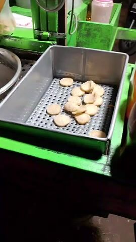 A person's hands are using tongs to move small, round, light-brown food items from one side of a perforated metal tray to the other. The tray is inside a larger green metal container, which appears to be part of a food cart. In the background, there are other food cart elements, including a pink bottle and some green tubing. The lighting suggests it is nighttime.