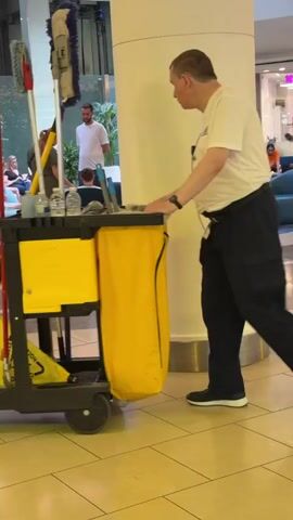 A man in a white t-shirt and dark pants pushes a yellow cleaning cart through a well-lit indoor space, possibly a mall. The cart is equipped with mops and other cleaning supplies. Several people are visible in the background, some sitting and others walking. The floor is tiled in a light color. The man's action of pushing the cart suggests he is performing cleaning duties.