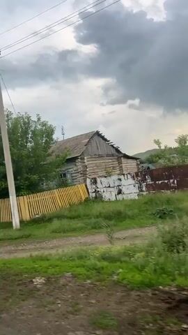 The video shows a roadside view of a rural village. A pedestrian crossing sign is visible on the left. A yellow wooden fence runs along the road, with a dilapidated wooden house behind it. Further down the road, there are more houses with blue fences. A speed bump sign and a speed limit sign of 40 are also visible. The sky is overcast with dark clouds. The road appears to be unpaved.