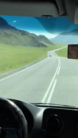 The video shows a view from the driver's seat of a vehicle, looking through the windshield. Outside, there is a paved road curving through a vast, green, mountainous landscape under a clear sky with a faint haze. The road has double white lines marking the lanes. A white car is visible in the distance on the road. The sun is shining brightly, casting shadows. The dashboard and steering wheel of the vehicle are visible in the foreground. A side mirror is attached to the windshield, reflecting the face of a person wearing glasses and a dark shirt. The overall scene depicts a scenic drive through a rural, possibly remote, mountainous region.