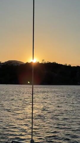 The video shows a view of a coastline at sunset from a boat. The sun is setting behind a hill with buildings on it. The camera pans from left to right, revealing more of the coastline, including houses, boats, a pier, and a large stone fortress on a hill. The water is choppy, and the sky is filled with light orange and pink clouds.