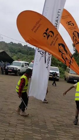 A group of people are gathered in an outdoor parking lot. Many are wearing bright vests, and some carry tall, curved flags with logos that seem to celebrate “25 Years of Geita Gold Mine.” The flags are in various colors, including orange and white. Some people are walking around while others appear to be dancing or moving rhythmically. There are several vehicles parked in the background, including vans and SUVs. The environment suggests a corporate event or celebration is taking place. The sky is overcast.