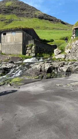 This media file showcases a scenic coastal landscape in Bøur, Faroe Islands. The scene transitions through different perspectives, beginning with a close-up of a small waterfall cascading over rocks. The water is clear and appears to be flowing into a dark, rocky area that leads towards a sandy shore.

The view then widens to reveal a building with a grass-covered roof, typical of Faroe Islands architecture, situated on a hillside. The building is constructed from concrete and stone, and the surrounding landscape is a vibrant green, suggesting a time of year with lush vegetation. The weather appears to be partly cloudy with bright sunlight.

Further shots reveal a black sand beach with waves gently lapping at the shore. In the distance, a dramatic, jagged mountain rises from the sea, its slopes covered in the same verdant green as the hillsides seen earlier. The overall atmosphere is serene and natural, highlighting the rugged beauty of the Faroe Islands. There is no visible text in the media file. The activity captured is primarily observational, appreciating the natural environment and the distinctive architecture.