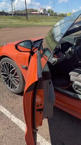 The video pans across the interior of an orange sports car. The driver's side door is open, revealing the gray and orange interior. The steering wheel, dashboard, and seats are visible. The car appears to be parked outdoors on a gravel surface, with a blurred background of a rural landscape, including buildings, a fence, and vegetation.