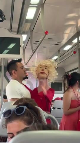 The video shows the interior of a public transport vehicle, likely a train or bus, with several passengers visible. In the foreground, the back of a woman's head with sunglasses on top of her hair is visible. In the middle ground, a man in a white t-shirt is standing and holding a puppet with blonde curly hair and an open mouth, wearing a red dress. The man's arm is around the puppet's shoulder. To the right of the puppet, a woman in a red dress is partially visible. The vehicle's interior features overhead lighting, handrails, and seats. The overall lighting suggests it is daytime. The puppet appears to be designed for performance or entertainment.