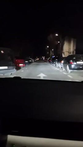 The camera is inside a vehicle driving at night on a street lined with parked cars on both sides. The street is dimly lit by streetlights. The vehicle's headlights illuminate the road ahead. There are no visible people or interactions. The focus is on the vehicle's perspective as it moves along the street.