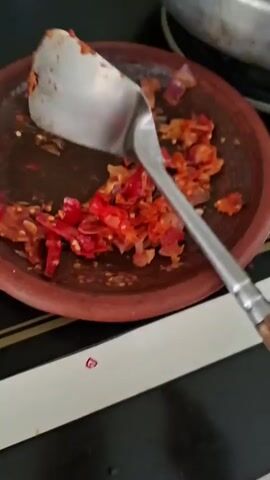 A close-up shot shows a dark brown clay mortar filled with chopped red chili peppers and onions. A silver spoon is used to stir the ingredients in the mortar. The mortar rests on a black surface with a white strip running along the bottom edge. In the background, a portion of a silver pot is visible. The lighting is dim, and the focus is on the contents of the mortar and the spoon.
