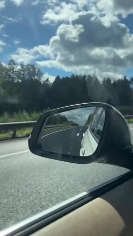 The video shows a view from inside a car, looking out the passenger side window and into the side mirror. The side mirror reflects a highway with white lane markings and a white car driving in the same direction. The sky is blue with fluffy white clouds, and the sun is shining. Trees and a guardrail are visible along the side of the highway. The interior of the car, including the door panel and window frame, is also visible in the foreground.