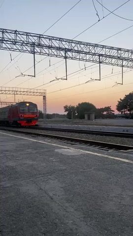 A red and gray train travels along railroad tracks at what appears to be dawn or dusk. The sky is a soft orange and pink. The train is passing a station platform with overhead electrical wires for the train. Trees are visible in the distance.