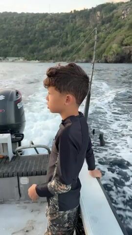 The video begins with a medium shot of a young boy standing on a boat. He is looking out to his left, away from the camera. He has dark hair and is wearing a black long-sleeved rash guard with camouflage-patterned shorts. Behind him to the left is the outboard motor of the boat, a black Suzuki with white lettering. To his right, a fishing rod with a reel is visible, extending upwards. The boat is in motion, leaving a white wake in the dark blue water. In the background, a lush green, hilly coastline is visible under a cloudy sky with hints of sunlight. The shot then pans slightly to the right, showing more of the boat's wake and the coastline. The boy turns his head slightly towards the camera, and his expression is neutral. The video continues to show the boat moving through the water, with the coastline remaining in the background. The camera then pans to the right again, focusing on the fishing rod and reel. The boy is still visible in the foreground, leaning on the edge of the boat. The video ends with a shot from behind the boy, looking out at the wake and the coastline.