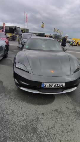 The video shows a parking lot with several Porsche Taycan Cross Turismo vehicles parked in rows. The cars are various shades of gray and silver. The environment suggests an event or gathering, with tents, flags, and equipment trailers visible in the background. People are walking in the background, but the focus is on the cars.