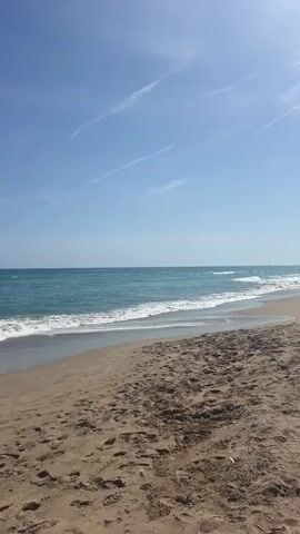 The video shows a beach on a sunny day. The sky is blue with a few wispy clouds. The ocean is turquoise and there are small waves breaking on the sandy shore. The sand is light brown and is covered in footprints. A single sailboat can be seen in the distance.