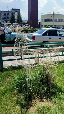 The video shows a wireframe deer statue in a grassy area. Behind the statue, there is a sidewalk and a street with several cars. In the background, there are buildings, including a tall, dark tower and a white, rectangular building. The sky is blue with some clouds. The overall impression is a public outdoor space, possibly a park or a roadside area, decorated with the deer statue.