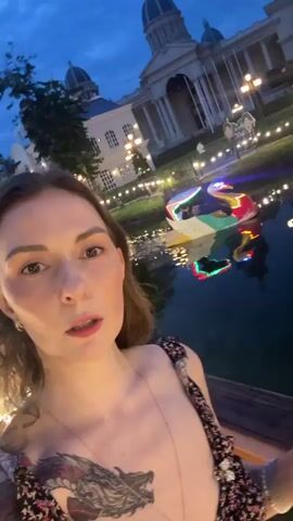 A woman with brown hair and a tattoo of a dragon on her chest films herself in front of a large, ornate building with a domed roof. She is wearing a dark floral-patterned dress and a necklace. The building is illuminated by lights and reflected in a body of water in front of it. A swan-shaped boat with colorful lights is visible in the water. The sky is dark, indicating it is either dusk or nighttime. The woman holds a small black object in her right hand.