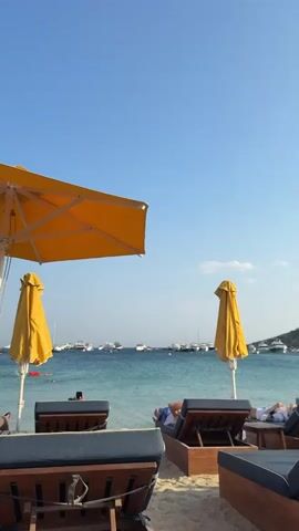 A beach scene with two yellow beach umbrellas, one open and partially visible in the foreground, and another closed and standing in the middle ground. In front of the umbrellas, there are several lounge chairs facing the ocean. The ocean is a clear turquoise color and dotted with numerous white boats. A few people are visible in the water, swimming. The sky is a clear, bright blue.