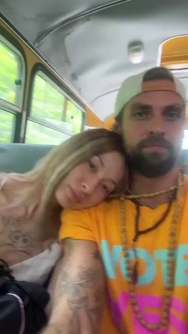 A couple is sitting inside a bus. The woman is leaning her head on the man's shoulder. Both are looking at the camera. The man is wearing a yellow t-shirt and a beige baseball cap. He has a beard and is wearing several necklaces. The woman has long blonde hair and is wearing a white top with a floral pattern. Her arms and chest are tattooed. The bus interior is visible with yellow accents on the ceiling and windows that show trees outside. The video appears to be a selfie video recorded by the man.