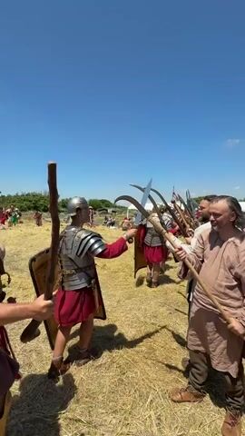 A video shows a re-enactment of a historical battle or event. Three men in the foreground are engaged in a mock fight. The man on the left is dressed as a Roman soldier, wearing a silver helmet, armor, and a red and white skirt. He holds a sword in his right hand. The man in the middle is also dressed as a Roman soldier, with a helmet, armor, and a red and white skirt, holding a sword. The man on the right is dressed in rustic clothing, a light brown tunic, and holds a long pole with a curved blade at the end, resembling a scythe or billhook. Other individuals in similar costumes are visible in the background, lined up as if preparing for battle. The scene takes place outdoors on a sunny day, with dry grass on the ground and a clear blue sky.