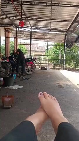A person's feet are in the foreground, with toenails painted dark purple. In the background, a person in a black shirt and pants is working on a motorcycle. The scene appears to be an outdoor covered area, possibly a repair shop or garage, with a concrete floor. There are tools and buckets visible on the ground near the motorcycle. Trees and buildings are visible in the distance.