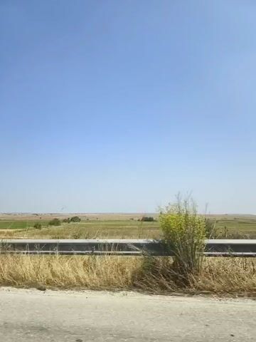 The video shows a view from a moving vehicle, likely a car, looking out at a landscape. The sky is clear and blue, occupying the upper portion of the frame. Below the sky is a flat, open field or plain. A guardrail made of metal poles and horizontal bars runs across the middle of the frame, separating the foreground from the background. The foreground consists of dry, yellowish grass and a grey, paved surface, likely a road. In the distance, the landscape appears to be agricultural fields, some green and some dry, with scattered trees or bushes. The overall impression is of a rural or semi-rural setting under a bright, sunny sky.