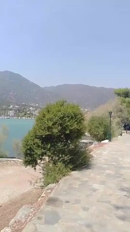 The video pans from a view of a bay with sailboats and hills in the background to a stone path leading up to a small, ornate church. The church has a red tiled roof, brick walls, and arched entrances with white columns. A Greek flag is flying on a flagpole in front of the church. There is a bell tower to the right of the church with another Greek flag. The sky is clear and blue. The environment is outdoors, with lush green trees and vegetation surrounding the path and church. The ground is paved with irregular stone slabs.