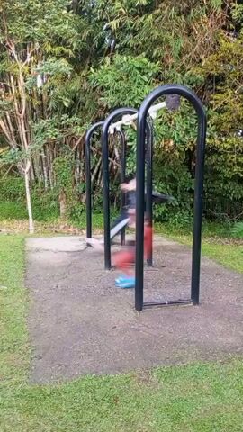 Two children are swinging on a playground set in an outdoor park. The child on the left is wearing a gray hooded jacket and dark pants. The child on the right is wearing a red shirt and dark shorts. The playground equipment consists of three black metal arch frames with horizontal bars at the top. The ground is a mixture of concrete and grass, surrounded by trees and foliage. The lighting suggests it is daytime.
