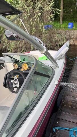 The camera, from a high angle, captures a man standing in a partially-covered boat docked on a wooden pier. The boat is predominantly white with a purple accent line along the hull. It features a steering wheel with a metallic finish, several circular gauges, and a windscreen. A silver metal frame structure, possibly a wakeboard tower, extends above the boat. The man is wearing a light-colored shirt and khaki pants.  Behind the boat, dense green foliage and glimpses of blue plastic containers are visible on land. A blue rope secures the boat to the pier. The camera appears to be handheld, resulting in slight movements. A conversation in Russian is audible.
