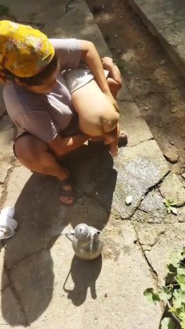 A woman wearing a headscarf and a t-shirt is squatting on a concrete path outdoors. She is holding a baby, whose lower body is exposed. The woman is pouring water from a metal pitcher onto the baby's diaper area. There is a shadow of the woman and pitcher on the ground. Some greenery is visible to the right.