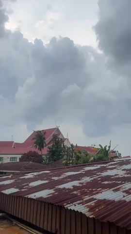 The video captures a view of rooftops and buildings under a cloudy sky. In the foreground, a corrugated metal roof, appearing aged and rusty, extends across the bottom of the frame. Behind this, a larger building with a red roof and light-colored walls is visible, surrounded by various trees and vegetation. The sky is filled with large, grey clouds, suggesting an overcast or possibly stormy weather condition. The overall scene is static, with no visible movement of people or distinct actions occurring. The lighting is diffused due to the cloud cover.