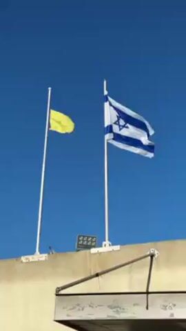 The video shows two flags, one yellow and one of Israel, waving on top of a beige building against a clear blue sky. Both flags are attached to white poles. There is a solar panel and a black cable visible on the roof of the building.